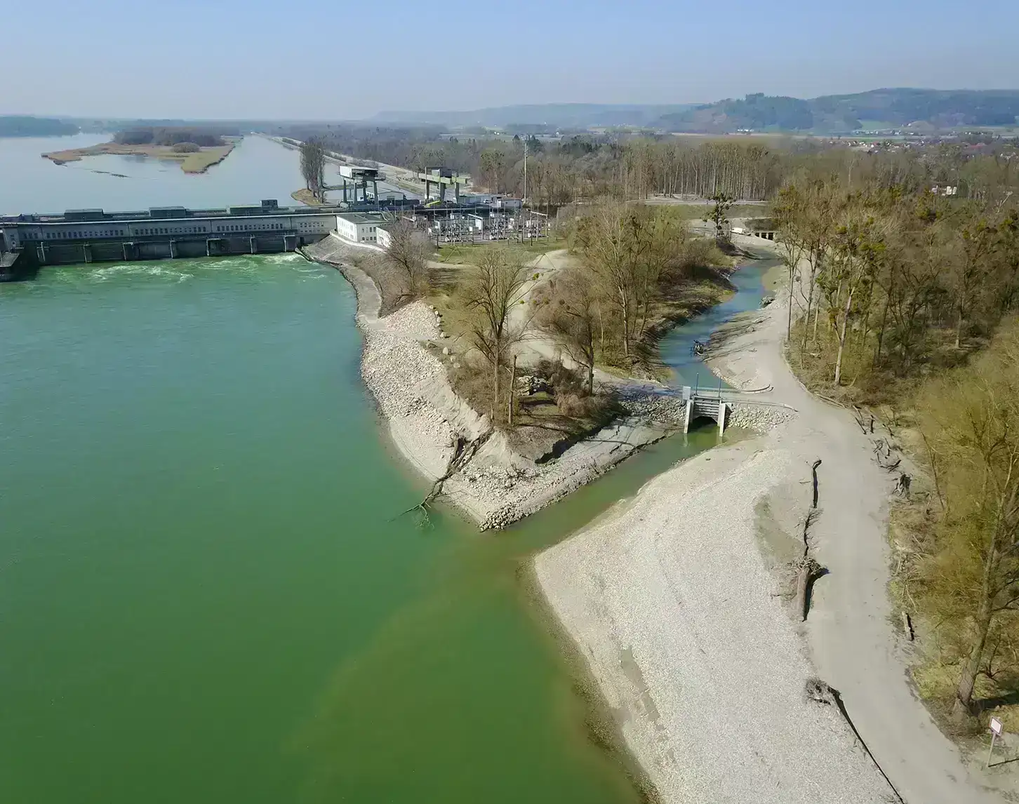 Aerial view of a power plant on a river with backed-up water, adjacent wooded area, and hiking trails in clear weather.