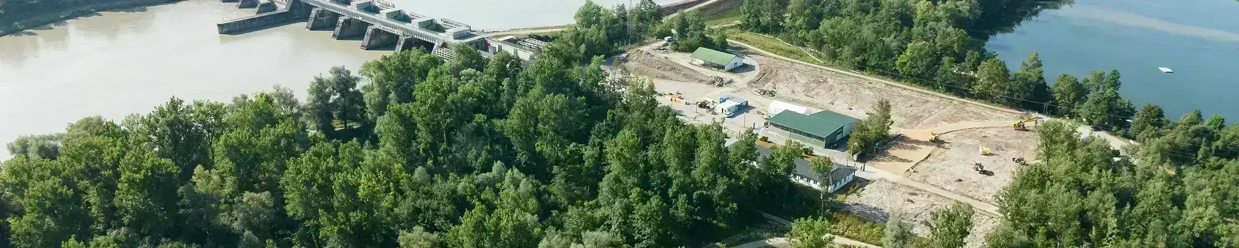 Luftaufnahme eines Wasserkraftwerks am Fluss mit einem Waldgebiet im Vordergrund und landwirtschaftlichen Feldern im Hintergrund.