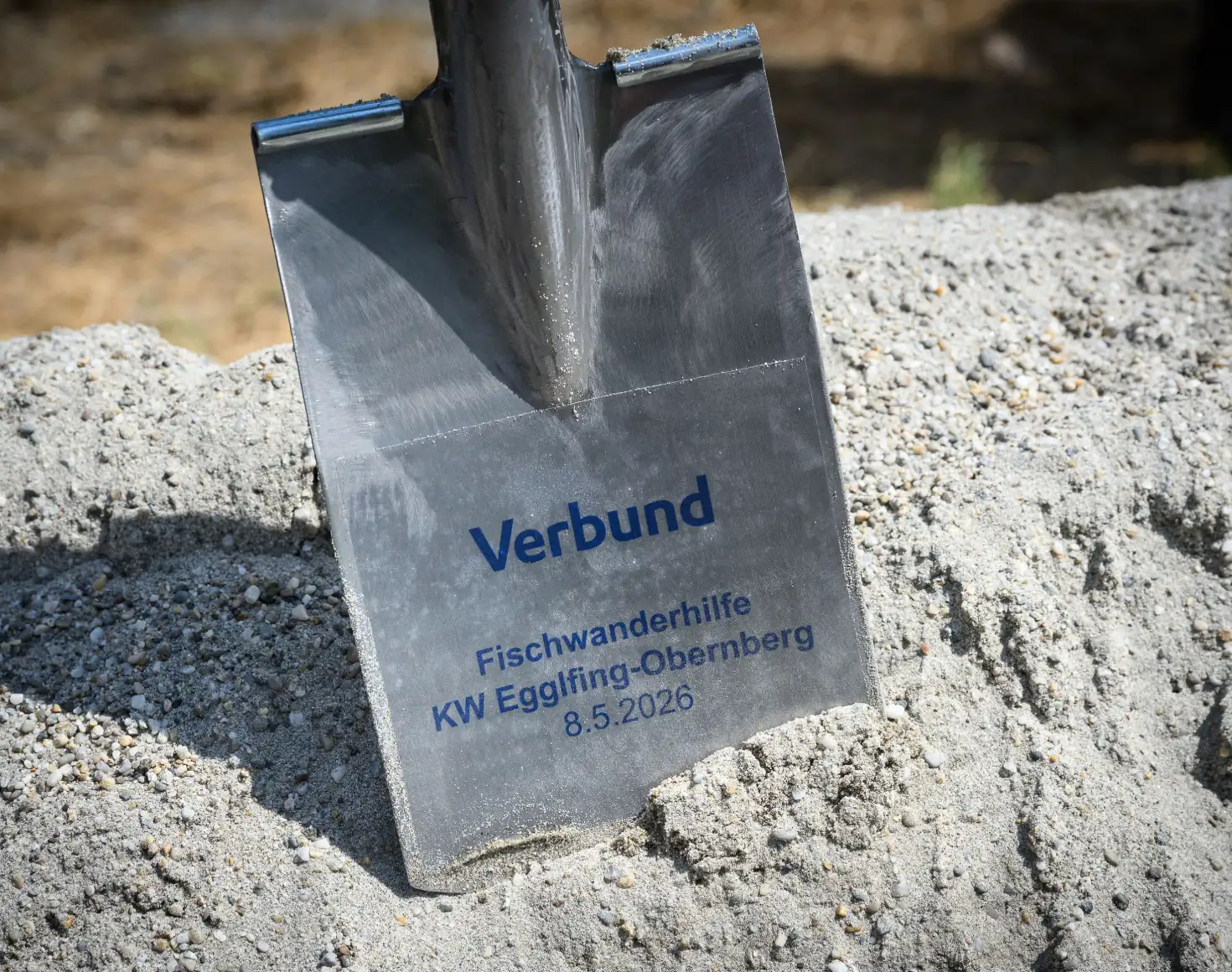 Shovel with engraved inscription “Verbund – Fish Passage KW Egglfing-Obernberg 8.5.2026” stuck in a pile of sand during the groundbreaking ceremony for the fish passage at the Egglfing-Obernberg power plant.