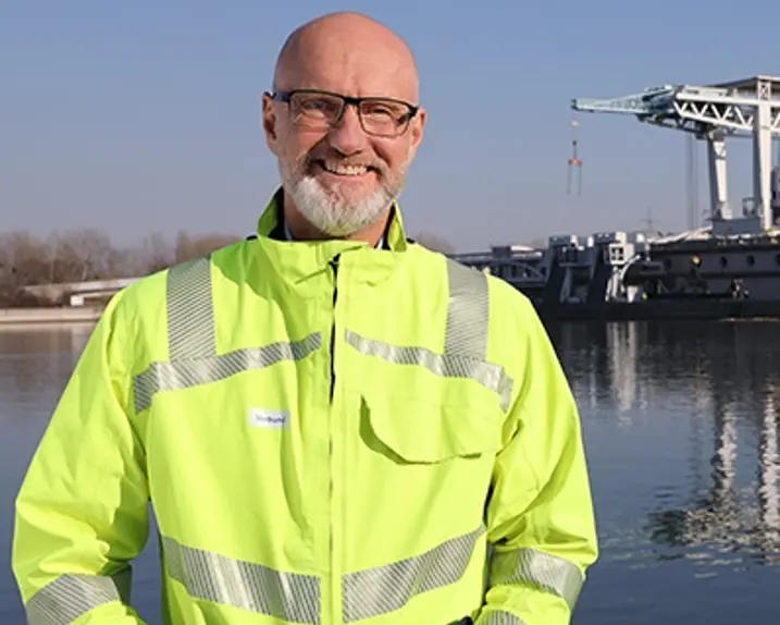 Rainer Tschopp trägt eine Schutzjacke in grellem gelb mit silbernen Reflektorstreifen. Er hat außerdem eine Brille und steht vor einem Kraftwerk bei Sonnenschein und lächelt in die Kamera.